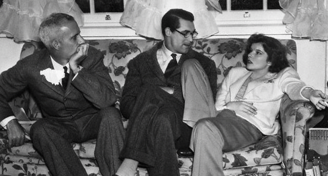 Howard Hawkes, Cary Grant and Katharine Hepburn sit on a floral patterned coach. Hawkes and Grant are looking at and listening to Hepburn as she slouches with one leg up on the couch, another touching the floor. She's wearing slacks and a sweater.