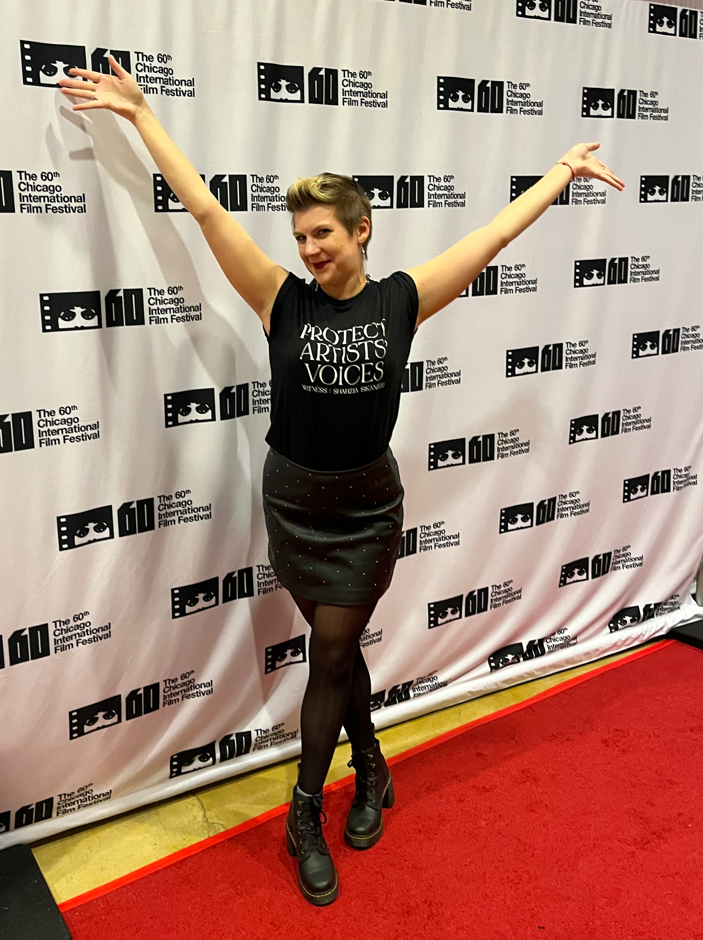 I'm a millennial white woman with a blond pixie haircut standing with my arms over my head on a red carpet. Behind me is a backdrop that says "The 60th Chicago International Film Festival." I'm wearing a black shirt that says "Protect Artists' Voices" and a black leather skirt with boots.